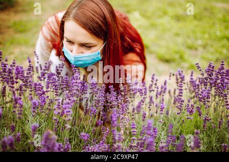 Jeune femme aux cheveux longs rouges portant un masque chirurgical essayant de sentir des fleurs de lavande pourpre. Allergie, covid, grippe Banque D'Images