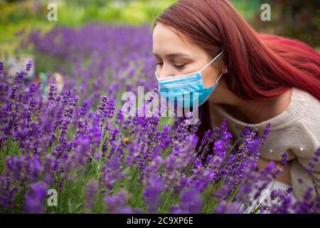 Jeune femme aux cheveux longs rouges portant un masque chirurgical essayant de sentir des fleurs de lavande pourpre. Allergie, covid, grippe Banque D'Images