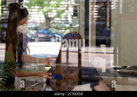 Jeune coiffeur avec masque facial peignant son client. Photographie avec réflexions de la fenêtre de la boutique. Nouvelle normalité. Banque D'Images