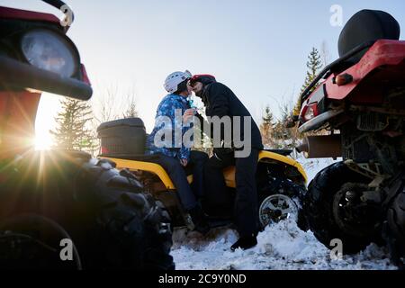 Couple amoureux assis sur un véhicule tout-terrain jaune et embrassant. Jeune femme et homme en casque tenant des verres de champagne et partageant le moment d'appel d'offres le jour d'hiver. Concept de l'amour et du quad. Banque D'Images