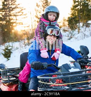 Portrait de famille, de père et de fille souriants, portant des costumes de ski, des casques et des lunettes de protection, assis à vélo d'équipe, souriant à la caméra dans des montagnes enneigées au coucher du soleil. Concept de loisirs actifs. Banque D'Images