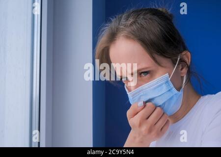 femme portant un masque, se sentant malade, souffrant de toux, regardant par la fenêtre dans la chambre avec le mur bleu - gros plan. Auto-isolation, prévention Banque D'Images