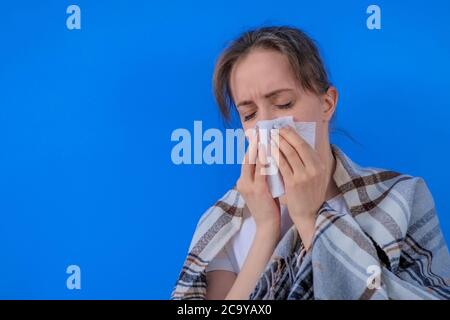 Femme portant une couverture à carreaux, se sentant malade, éternuant dans un mouchoir en papier dans la chambre avec un mur bleu à la maison. Santé, espace de copie, quarantaine, maladie Banque D'Images