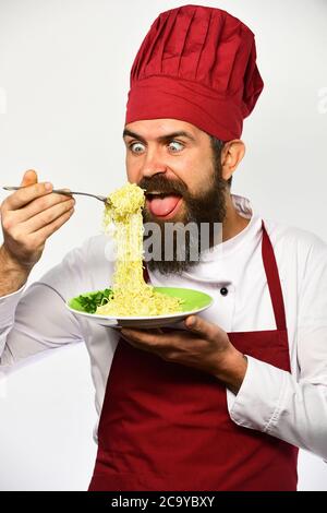 L'homme avec la barbe tient un plat savoureux sur fond blanc. Cuire avec le visage affamé en uniforme bordeaux contient la fourchette et l'assiette. Le chef mange des nouilles italiennes ou asiatiques. Concept de restauration rapide. Banque D'Images