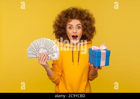 Ouah, cashback, shopping ! Portrait d'une femme surprise à cheveux maussés dans un sweat à capuche de style urbain tenant de l'argent, des billets en dollars et une boîte cadeau, expressin Banque D'Images