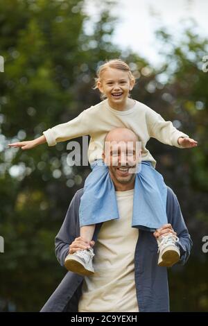 Portrait vertical d'une jolie fille assise sur les épaules des papas et s'amusant tout en appréciant la marche dans le parc à l'extérieur, les deux souriant à l'appareil photo Banque D'Images