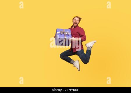 Enthousiaste extrêmement joyeux hipster gars sautant trampoline avec boîte cadeau, volant dans l'air tenant anniversaire présent surprise, appréciant bonus de vacances. f Banque D'Images