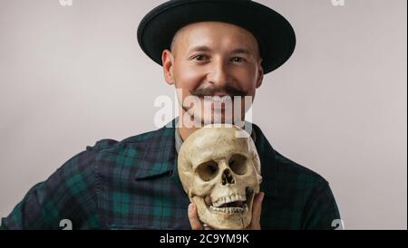 Cute man wearing hat holding élégant crâne humain dans la main et à la caméra à plus de studio fond blanc Banque D'Images