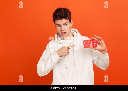 Surprise jeune homme en blanc sweat-shirt avec capuche tenant la carte de réduction dans la main choqué avec méga vente, shopaholic. Studio d'intérieur tourné isolé sur Ora Banque D'Images