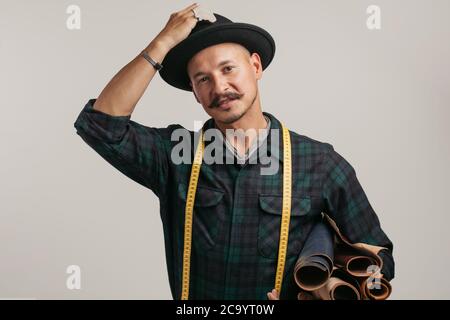 Mignon caucasien joyeux moustached handyman dans le chapeau créatif, posant avec des rouleaux de cuir isolés sur fond gris studio Banque D'Images