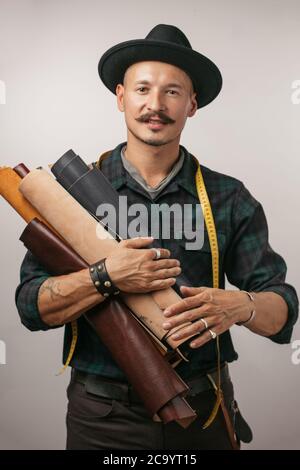 Mignon caucasien joyeux moustached handyman dans le chapeau créatif, posant avec des rouleaux de cuir isolés sur fond gris studio Banque D'Images