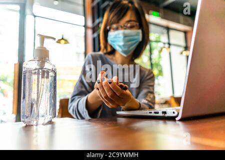 Jeune femme se lavant les mains avec du gel d'alcool en quarantaine pour le coronavirus portant un masque de protection avec distanciation sociale et utilisant le travail d'ordinateur portable Banque D'Images