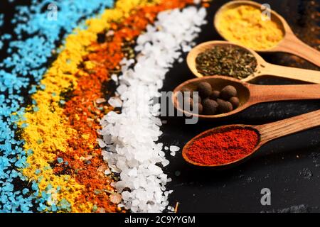 Concept de l'art alimentaire. Cuillères en bois avec paprika, herbes, curcuma et boules de poivre près de sel de mer granulé. Ensemble d'épices sur fond de bois gris foncé. Composition de sel bleu, blanc, orange et jaune Banque D'Images