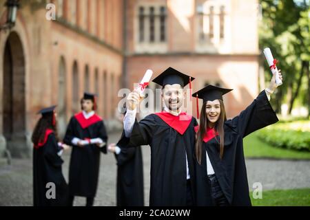 Deux diplômés enthousiastes en robes debout et montrant le certificat devant les étudiants Banque D'Images