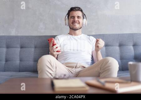 Profiter de sa musique préférée. Un jeune homme plein de gaieté, dans un casque, qui écoute la musique tout en se assis sur son canapé à la maison. Banque D'Images