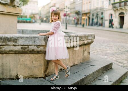 Adorable petite fille blonde dans une belle robe rose posant à l'appareil photo à la vieille fontaine de la ville européenne antique en plein air. Portrait complet de Happy