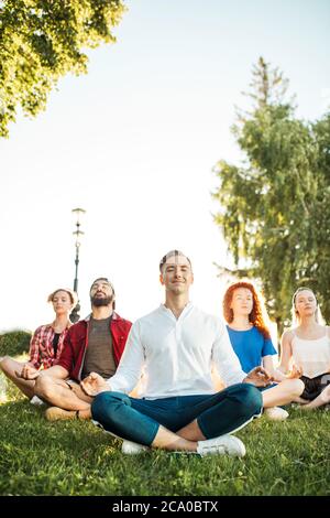 Divers cours de yoga de jeunes amis gaies en été vêtements décontractés assis dans lotus poser sur vert lown dans le parc public, vue de face avec accent sur ma Banque D'Images