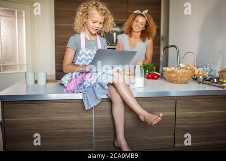 Adorable petite fille avec un frisé blond et sa mère cally aux cheveux justes avec la cuisine ensemble à la maison. Une fille qui lit la recette sur un ordinateur portable Banque D'Images