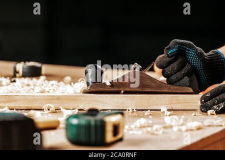 Gros plan du constructeur d'un jeune homme dans des clothers de travail et des gants noirs traite une barre en bois avec un avion de cric dans l'atelier Banque D'Images