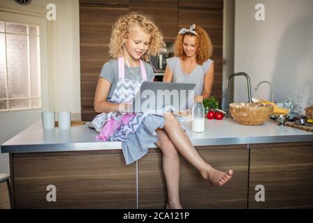 Adorable petite fille avec un frisé blond et sa mère cally aux cheveux justes avec la cuisine ensemble à la maison. Une fille qui lit la recette sur un ordinateur portable Banque D'Images