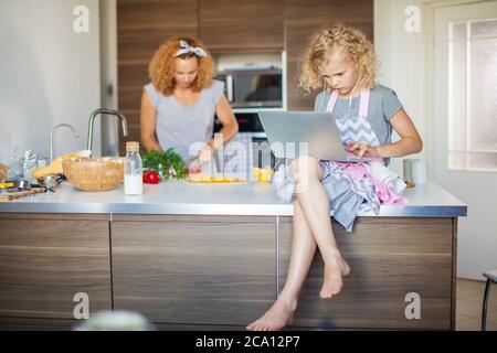 Portrait d'une femme au foyer caucasienne heureuse avec un dîner de cuisine de cheveux crépus à la maison avec sa fille d'âge préscolaire allongé sur une table avec un ordinateur portable. Banque D'Images