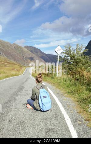 Une jeune fille assise sur la route asphaltée bénéficiant de la vue magnifique et de la liberté à Glencoe dans les Highlands écossais en été, en Écosse, en Grande-Bretagne Banque D'Images