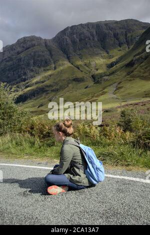 Une jeune fille assise sur la route asphaltée bénéficiant de la vue magnifique et de la liberté à Glencoe dans les Highlands écossais en été, en Écosse, en Grande-Bretagne Banque D'Images