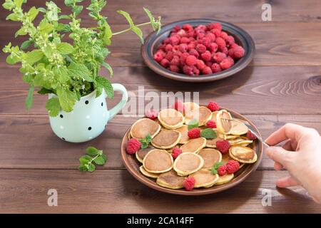 La main avec la fourchette atteint pour les céréales Pancake. Framboises fraîches mûres rouges dans une assiette brune et un bouquet de Melissa dans une tasse sur un t en bois foncé Banque D'Images