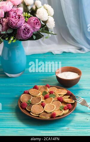 Céréales pancakes et framboises fraîches mûres rouges dans une assiette brune. Un bouquet de pivoines dans un vase sur une table en bois bleu près de la fenêtre. Délicieux summ Banque D'Images