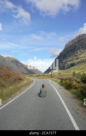 Une jeune fille assise sur la route asphaltée bénéficiant de la vue magnifique et de la liberté à Glencoe dans les Highlands écossais en été, en Écosse, en Grande-Bretagne Banque D'Images