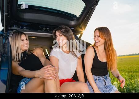 Les jeunes femmes blonde, brune et aux cheveux rouges s'assoient et se détendent au dos du véhicule, parlant lors d'un voyage aventure en nature. Banque D'Images