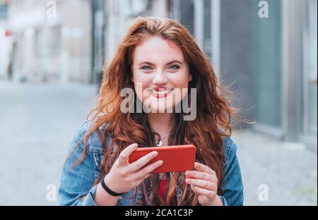 Portrait de sourire rouge mauridé cheveux longs caucasienne jeune fille marchant dans la rue et chat vidéo à l'aide du smartphone moderne. Les gens modernes avec Banque D'Images