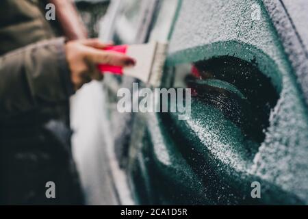 Une femme nettoie une fenêtre glacée sur une voiture avec un grattoir à glace. Concentrez-vous sur la glace de la fenêtre. Matin enneigé et gelé. Banque D'Images