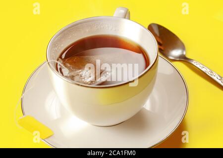 Tasse en porcelaine blanche avec thé chaud fraîchement préparé, sac encore dans, cuillère en argent à côté sur le tableau jaune, éclairé par le soleil du matin Banque D'Images