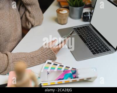 Photo courte d'une femme de créateur dans un chandail à l'aide d'un smartphone tout en travaillant avec un ordinateur portable factice sur le bureau Banque D'Images