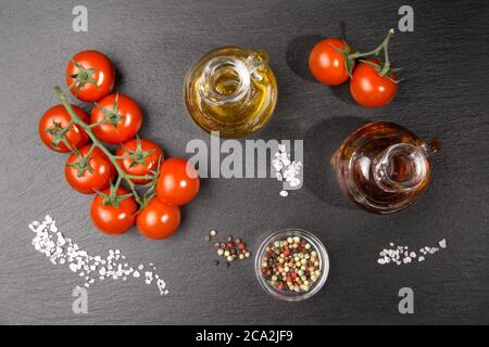 Une brindille de tomates cerises avec quelques grains de sel épais, une huile et une carafe de vinaigre et du poivre coloré sur une dalle d'ardoise. Banque D'Images