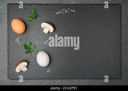 Omelette aux champignons avec deux champignons demi-bruns, un œuf brun et un œuf blanc, deux feuilles de persil et de gros grains de sel sur une plaque d'ardoise. Avec co Banque D'Images