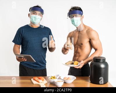 deux blogueurs asiatiques de santé et de sport portant un masque et écran facial avec les pouces vers le haut montrant le menu des aliments sains pendant tenez un support de tablette orienté vers l'avant et regardez l'appareil photo sur un arrière-plan isolé Banque D'Images