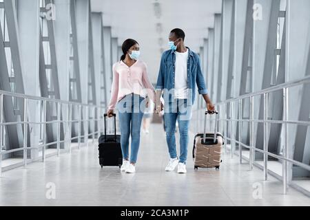 Le coronovirus se déplace. Couple noir dans des masques marchant avec des bagages dans le bâtiment de l'aéroport Banque D'Images