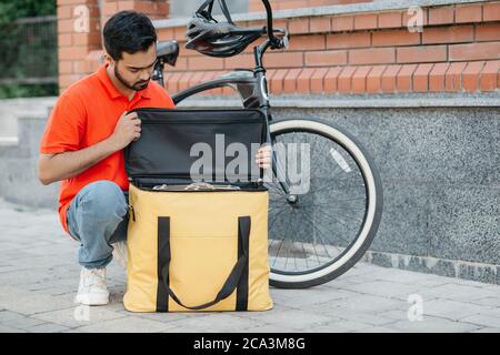 Emballage de la commande dans un sac thermique. Un vrai messager de type avec barbe en uniforme avec un vélo regarde le sac jaune Banque D'Images