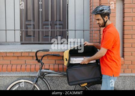 Livraison à domicile et au bureau. Un jeune livreur prend une pizza dans un sac à dos jaune sur un vélo Banque D'Images