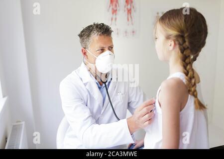 un bon médecin de sexe masculin avec une barbe de trois jours et un masque de nez et de bouche examine une jeune fille avec un stéthoscope Banque D'Images