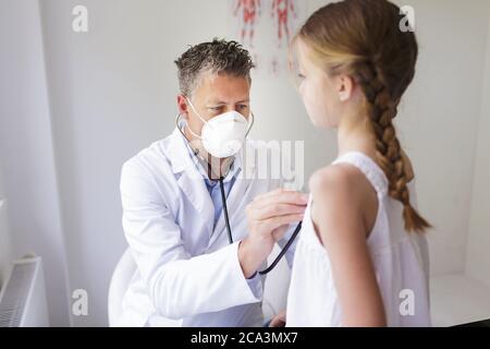 un bon médecin de sexe masculin avec une barbe de trois jours et un masque de nez et de bouche examine une jeune fille avec un stéthoscope Banque D'Images