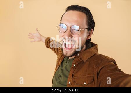 Image d'un jeune homme ravi qui se fait rire et se fait rire tout en prenant une photo de selfie isolée sur fond beige Banque D'Images