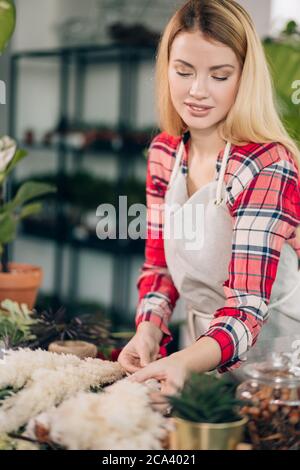 femme de fleuriste soigneuse avec de longs cheveux blonds en rouge décontracté damier chemise et tablier dans son propre magasin de fleuriste, s'occuper des plantes et des fleurs Banque D'Images