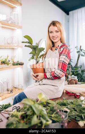 portrait d'une belle femme floriste caucasienne attrayante aux cheveux blonds, portant un tablier blanc sur une chemise à carreaux rouges et tenant des plantes dans des pots, surro Banque D'Images