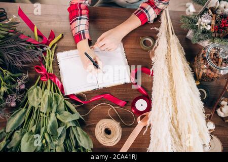 vue de dessus sur une jeune fleuriste femme prenant des notes au travail, ruban rouge, carnet et fleurs couchés sur une table. Concept de gens, d'affaires, de vente et de fleuriste Banque D'Images