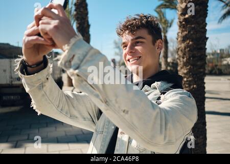 Portrait d'un homme souriant et attrayant prenant des photos avec joie sur un smartphone en extérieur Banque D'Images