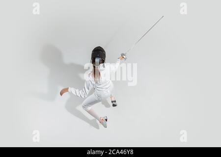 D'abord. Jeune fille en costume d'escrime avec épée à la main sur fond blanc. Vue de dessus. Jeune femme modèle pratique et formation en mouvement, action. CopySpace. Sport, jeunesse, mode de vie sain. Banque D'Images