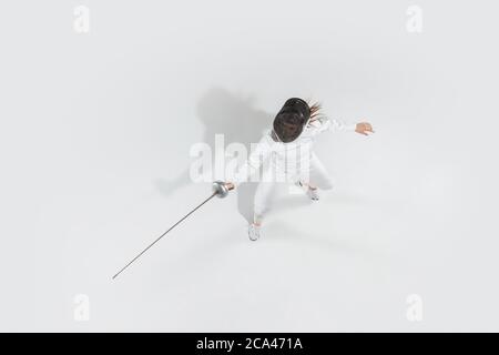 D'abord. Jeune fille en costume d'escrime avec épée à la main sur fond blanc. Vue de dessus. Jeune femme modèle pratique et formation en mouvement, action. CopySpace. Sport, jeunesse, mode de vie sain. Banque D'Images